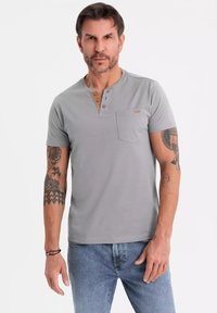 Camisa henley gris de manga corta con tres botones de madera, que presenta un bolsillo en el pecho y una textura suave y lisa. Llevada con jeans azules.