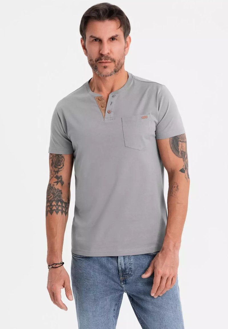 Camisa henley gris de manga corta con tres botones de madera, que presenta un bolsillo en el pecho y una textura suave y lisa. Llevada con jeans azules.