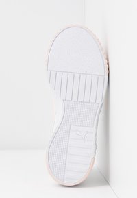 Bílá podrážka tenisek s texturovanými gripovými vzory a logem Puma, umístěná vzpřímeně proti bílým rohovým plochám.