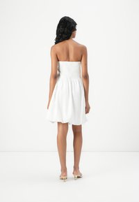 Hvid strapless kjole med smocked liv og udvidet nederdel. Glat tekstur, knælængde, båret med neutrale åben-tå hæle.