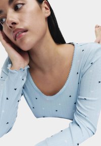 Lichtblauw shirt met lange mouwen en een ronde hals, met kleine zwarte en witte hartjespatronen en golvende randen aan de mouwen.