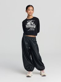 Czarny krótki top z długim rękawem z białym nadrukiem graficznym, zestawiony z luźnymi czarnymi spodniami z imitacji skóry oraz jasnymi sneakersami.