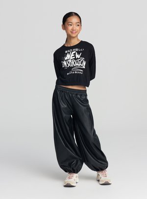 Crop top nero a maniche lunghe con stampa grafica bianca, abbinato a pantaloni larghi neri in ecopelle e sneakers di colore chiaro.