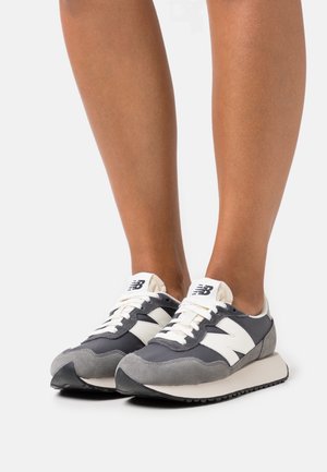 Nogi w szaro-białych sneakersach New Balance z białymi sznurówkami, stojące na białej powierzchni na jednolitym tle.