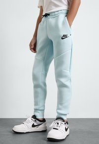 Šviesiai mėlynos, siauro kirpimo Nike sportinės kelnės su juodu logotipu. Yra elastinė juosmens juosta su raišteliu ir šoninės kišenės. Nešiojamos su baltomis sportbačiais.