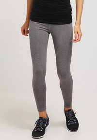 Personne portant des leggings gris ajustés, des chaussures de sport noires avec des semelles blanches et un haut noir, se tenant sur un fond clair.