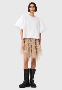 Top corto in cotone bianco con maniche a sbuffo, gonna di tulle beige a strati con balze e stivali neri in pelle al ginocchio con fibbia.