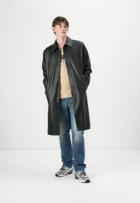 Schwarzer Kunstledermantel mit Kragen, Knöpfen auf der Vorderseite, kombiniert mit einem beigen Hemd und blauen Jeans. Das Model trägt schwarz-weiße Turnschuhe.