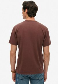 Mężczyzna z krótkimi ciemnymi włosami, ubrany w jednolitą brązową koszulkę z krótkim rękawem i niebieskie dżinsy, widoczny od tyłu na białym tle.