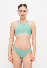 Smaragdzöld bikini szett, csillogó textúrával, sportos, magas nyakú top és alacsony derekú, varrat nélküli alsó rész.