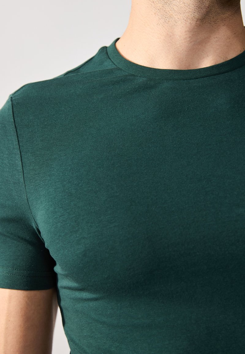 Zielona koszulka z krótkim rękawem wykonana z miękkiego materiału, z okrągłym dekoltem i gładką teksturą, z widocznymi detalami szwów.