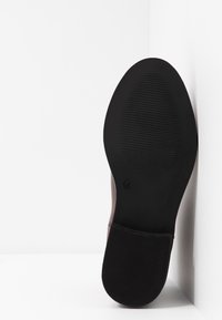 Suela de un zapato de cuero marrón oscuro con un talón de goma texturizada y agarre en la puntera, posicionada verticalmente contra una esquina blanca.