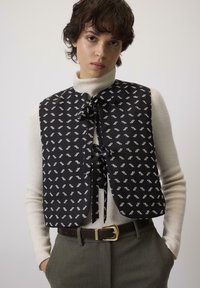 Crni prsluk s bijelim apstraktnim uzorkom, s vezanjem, obučen preko kreme rolke, u kombinaciji s sivim hlačama visokog struka i smešnim pojasom.