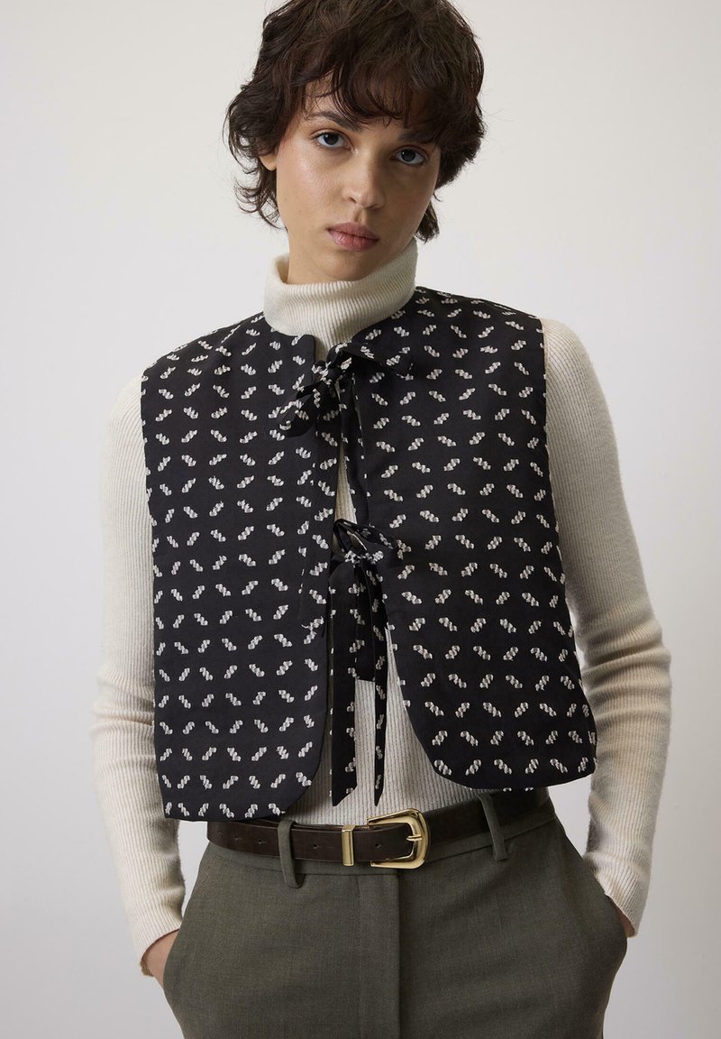 Crni prsluk s bijelim apstraktnim uzorkom, s vezanjem, obučen preko kreme rolke, u kombinaciji s sivim hlačama visokog struka i smešnim pojasom.