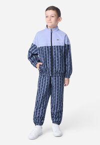 Otroški trenirni komplet v modri in mornarsko modri barvi z geometrijskim vzorcem. Značilnosti vključujejo visok ovratnik, zadrgo in elastične manšete. Bele športne copate.