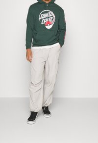 Hombre vistiendo una sudadera de Santa Cruz en verde oscuro con un gráfico de una Pokébola, pantalones cargo beige claros y deportivas negras de caña alta, de pie con las manos en los bolsillos.