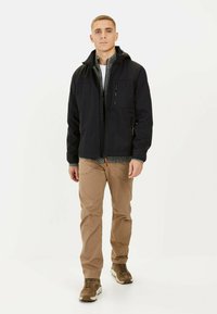 Veste zippée noire avec une doublure intérieure texturée, capuche et plusieurs poches, associée à un pantalon beige et des chaussures de sport marron.