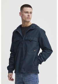 Mørkeblå, hettejakke med vindbryter design, to frontlommer og elastiske mansjetter, laget av teksturert stoff med et subtilt stripemønster.