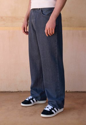 Person, die lockere dunkelgraue Jeans und schwarz-weiße Sneakers trägt, steht auf einem braunen Boden vor einer beigen Wand.