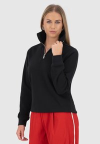 Pullover sweatshirt nero con zip a quarti, in morbido tessuto e polsini a coste, abbinato a pantaloni rossi vivaci con strisce bianche ai lati.