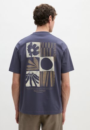 Mann trägt ein marineblaues T-Shirt mit abstrakten Blumen- und geometrischen Mustern und dem Schriftzug "Naturalness" auf der Rückseite, steht vor einem schlichten Hintergrund.