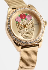 Gold mesh watch with a round face featuring a colorful sugar skull design, surrounded by a ring of clear crystals. Brand: Guess.