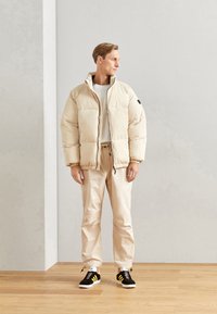Beige pufferjakke med høj krave, sidelommer og elastiske manchetter, båret over en lys grå skjorte og beige bukser, kombineret med sorte sneakers.