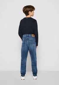 Jeans in denim blu con un lavaggio chiaro, tasche posteriori e un'etichetta marrone di Tommy Hilfiger. Abbinati a una maglietta a maniche lunghe di colore blu navy e sneakers bianche.