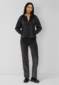 Junge Frau steht mit den Händen hinter dem Rücken, trägt eine schwarze Satin-Bluse mit langen Ärmeln, schwarze Hose und glänzende schwarze Schuhe.