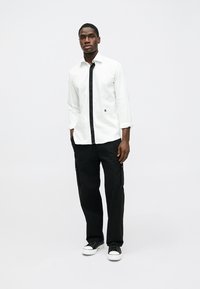 Camisa branca de manga comprida com uma pala frontal preta, combinada com calças cargo pretas e ténis brancos, apresentando linhas limpas e uma silhueta ajustada.