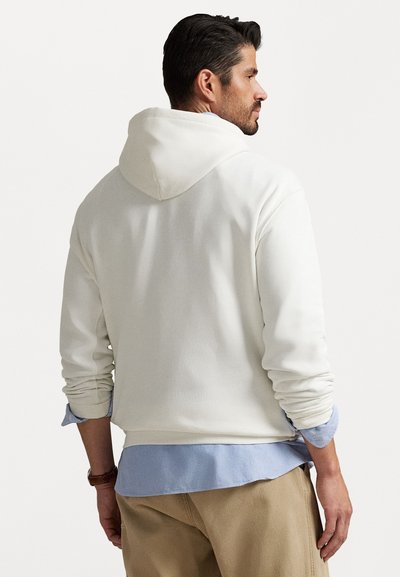 Homem a usar um hoodie off-white sobre uma camisa azul clara e calças beijas, de costas contra um fundo branco liso.
