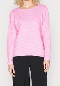 Hellrosa langärmliger Pullover mit strukturiertem, blasenartig gemustertem Stoff und rundem Ausschnitt, kombiniert mit schwarzen weit geschnittenen Hosen.