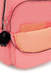 Zaino corallo rosa con un tessuto testurizzato, dotato di una tasca con zip e una morbida patta in tono pesca a contrasto. Dettagli in zip nera e charm.