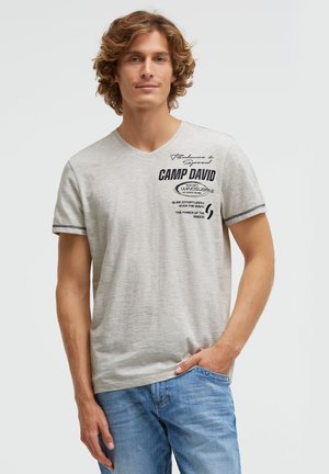 Young man with curly light brown hair wearing a light gray Camp David t-shirt and blue jeans, standing with one hand in pocket.