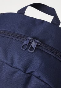 Navy rugzak met dubbele ritssluitingen, textuurstof en een handvat aan de bovenkant om te dragen. De ritskokers zijn blauw en gelabeld.