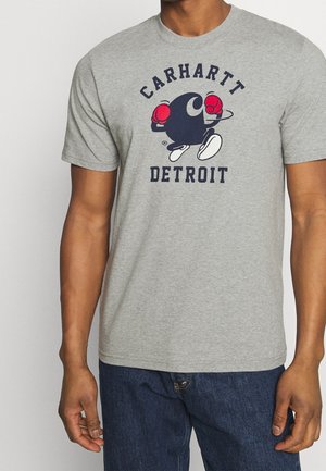 Persona que lleva una camiseta gris con un personaje de círculo negro de dibujos animados que lleva guantes de boxeo rojos y zapatos blancos, el texto dice Carhartt Detroit.