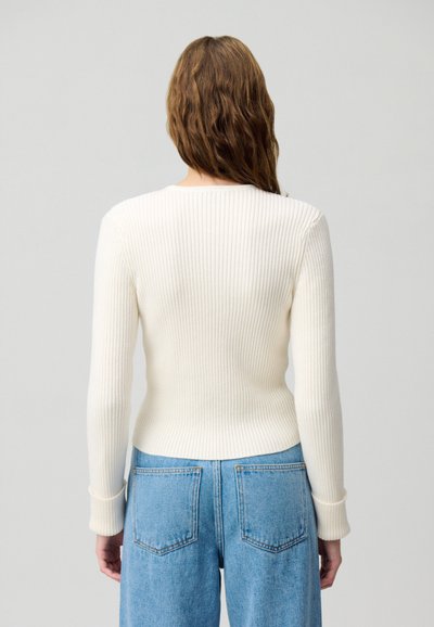 Top a maniche lunghe a coste color crema, con scollatura rotonda e silhouette aderente. Abbinato a pantaloni in denim blu a vita alta.