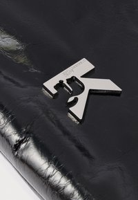 Spīdīga melna ādas virsma ar sudraba metāla "FK" logotipu un mazu iegravētu tekstu "KARL LAGERFELD JEANS" augšpusē.