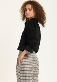 Black button-up shirt with rolled sleeves, paired with checkered high-waisted trousers. The fabric appears smooth, with a relaxed fit.