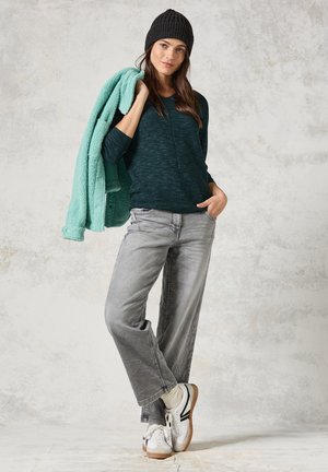 Donkerblauwe sweater met textuur, grijze wijde jeans en witte sneakers met zwarte accenten. Zachte teal fleece jas over de schouder gedrapeerd, zwarte gebreide beanie.