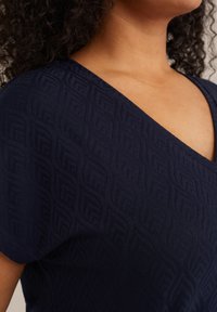 Mörkblå topp med ett strukturerat mönster och V-ringning. Tyget har subtila detaljer och en mjuk textur.