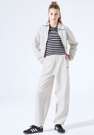 Junge Frau steht mit den Händen in den Taschen, trägt beige Jogginghose und Jacke, ein schwarz-weiß gestreiftes Shirt und schwarze Sneakers mit weißen Streifen.