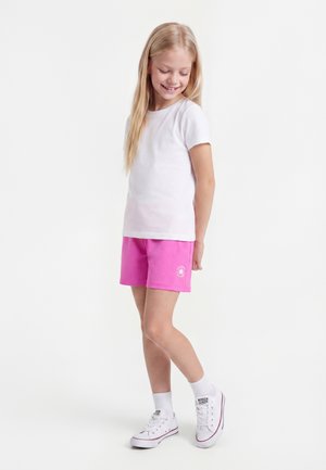 Jeune fille souriante, portant un t-shirt blanc, un short rose avec un logo, des chaussettes blanches et des baskets blanches, debout devant un fond blanc uni.
