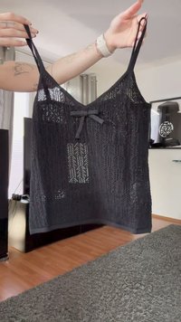 Händer som håller en svart spetscamisole med tunna remmar och en liten rosett i mitten, inomhus på ett trägolv med en grå matta.
