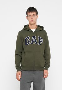 Zip-up dukserica u maslinastoj zeleni boji od pamuka, s velikim mornarsko plavim "GAP" logotipom, prednjim džepovima i kapuljačom s špagu. Glatka tekstura.