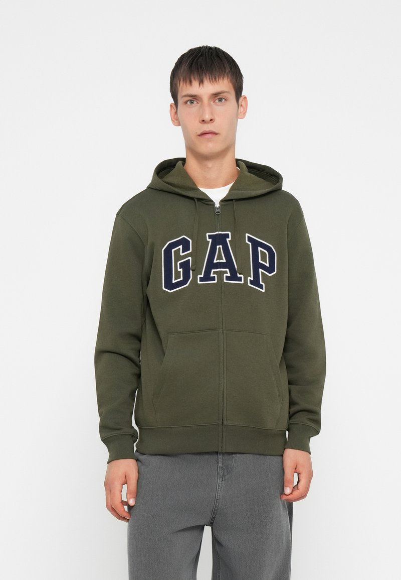 Zip-up dukserica u maslinastoj zeleni boji od pamuka, s velikim mornarsko plavim "GAP" logotipom, prednjim džepovima i kapuljačom s špagu. Glatka tekstura.