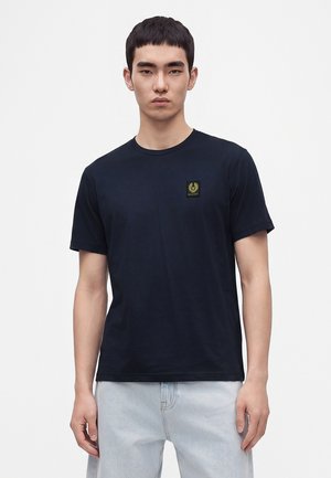 Giovane uomo che indossa una maglietta nera semplice a maniche corte con un piccolo logo sul petto e jeans azzurri, in piedi su sfondo bianco.
