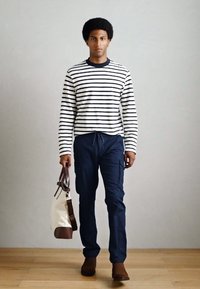 Stribet langærmet skjorte i hvid og marineblå, marineblå cargo-bukser, brune ankelstøvler og en cremefarvet taske med brune detaljer. Model går på trægulv.