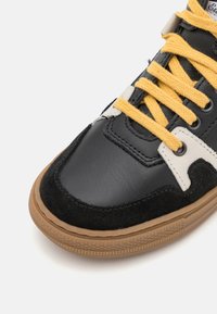 Zwarte leren en suède sneaker met gele veters, witte accenten en een gestructureerde gumrubberen zool. Kenmerkt zich door een ronde neus.
