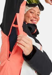Giacca da sci nera e corallo con apertura sottoascella con zip, caratterizzata da una striscia di design bianca e una fodera interna testurizzata.